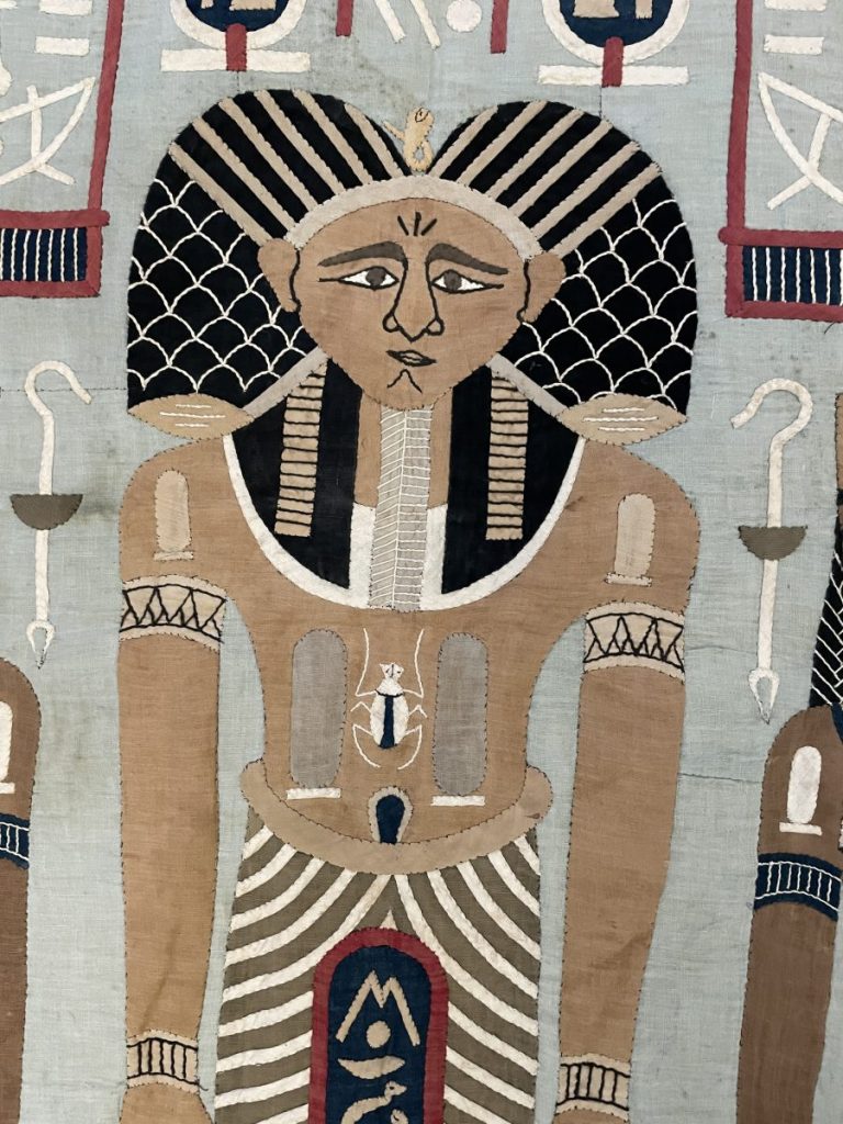 Textile artwork depicting an ancient Egyptian figure with elaborate headdress and detailed attire.
