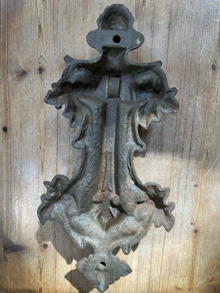 An ornate dark cast-iron wall ornament with floral relief and two mounting holes rests against weathered wood boards in bright daylight outdoors.
